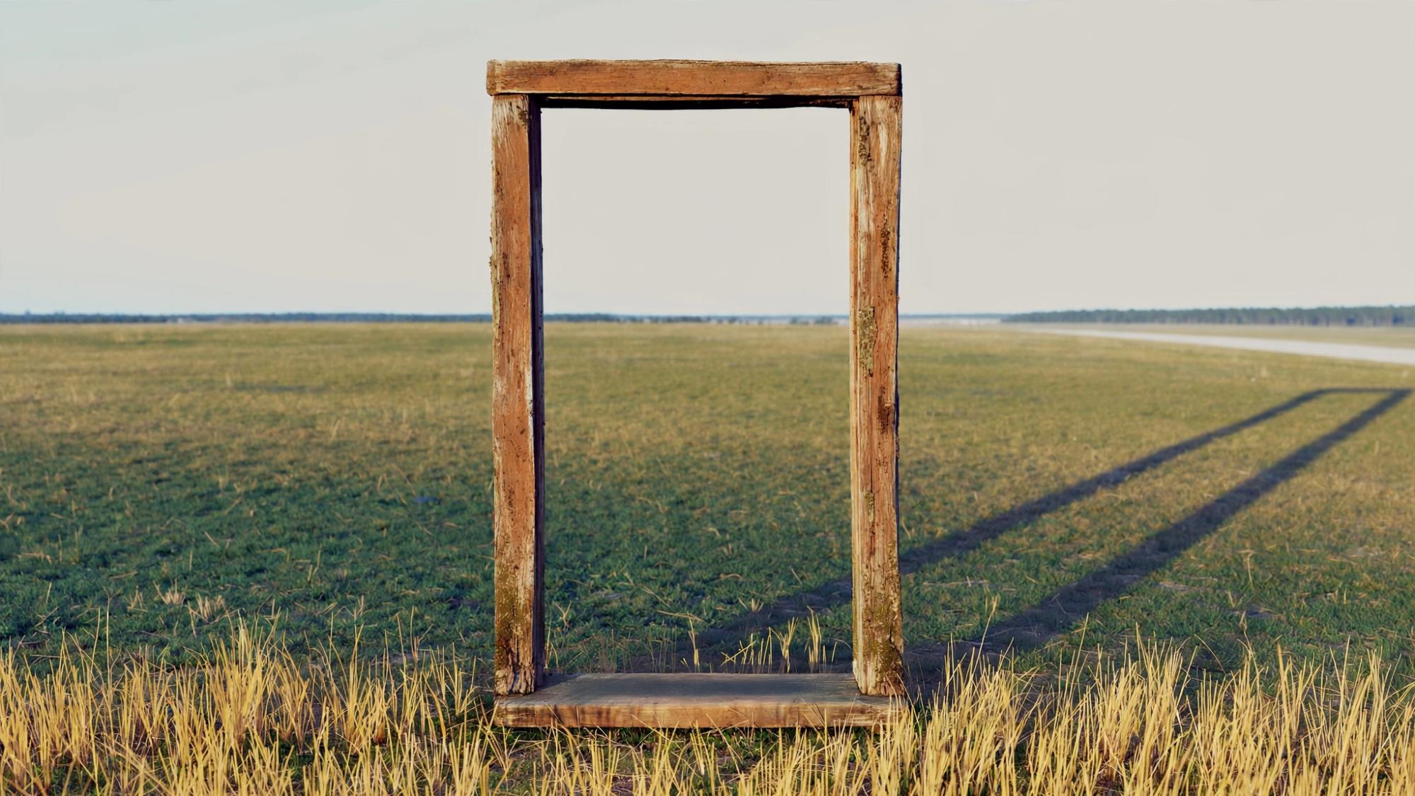 A wooden doorway standing alone in a field, its shadow long and pointed back toward the road.